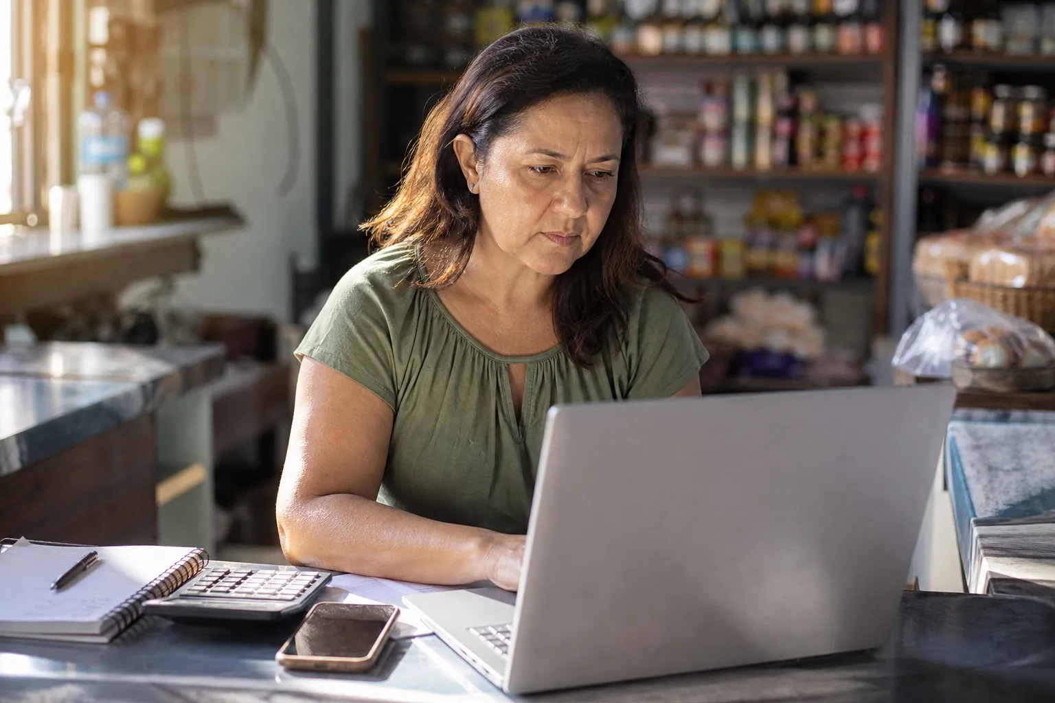 Dona de loja conferindo a fatura do mês no notebook, com calculadora e celular no balcão